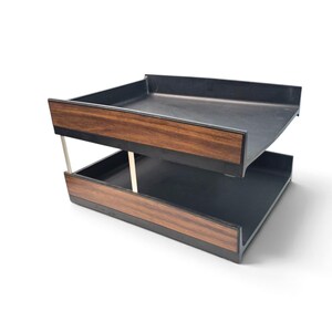 May include: A two-tier desk organizer featuring black plastic trays with a faux wood grain finish on the front panels. The trays are connected by thin, vertical silver-colored metal supports, creating a stacked structure for document storage.