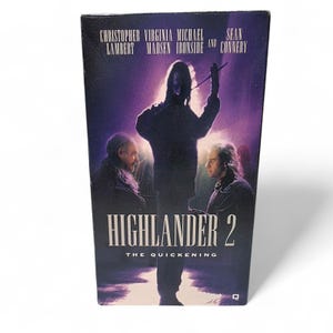 May include: A VHS tape for the movie "Highlander 2: The Quickening." The cover features a dark silhouette of a figure holding a staff, with other figures and the movie title in white. The tape is in a black case.