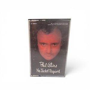 May include: A music cassette tape with a clear plastic case featuring the album cover for Phil Collins' No Jacket Required. The cover displays a close-up portrait of a person with a red-tinted face against a black background, with white text.
