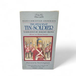 May include: A VHS video tape of "The Steadfast Tin Soldier" by Hans Christian Andersen. The cover features an illustration of toy soldiers in red coats and a central figure in a red uniform. The text includes the title, narrator, and illustrator.