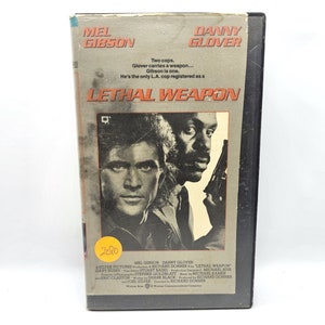 May include: VHS video cassette cover for the action film "Lethal Weapon" starring Mel Gibson and Danny Glover. The cover features a close-up of Gibson and Glover with guns in hand. The title "Lethal Weapon" is in large, bold letters.