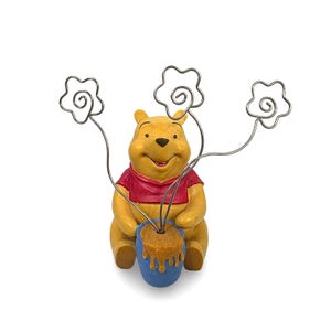 May include: A Winnie the Pooh figurine holding a blue honey pot with three silver wire flower-shaped holders. The figurine is yellow with a red shirt and a smiling expression. Honey drips from the pot.