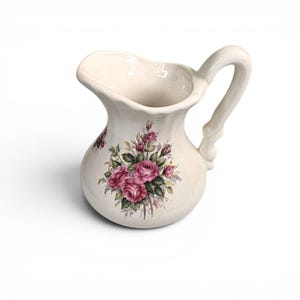 May include: Cream-colored ceramic pitcher with a floral rose design in shades of pink and green. The pitcher has a curved handle and a flared spout. A decorative piece for the kitchen or dining room.