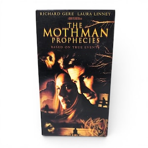 May include: A VHS tape case for the film The Mothman Prophecies, featuring actors Richard Gere and Laura Linney. The cover art is dark with orange and black tones, displaying the movie title, the text Based on True Events, and a Mark Pellington film.