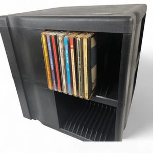 May include: A black plastic CD storage unit with multiple shelves. Several CD cases are visible, with colorful album art and text. The unit is designed to hold and organize compact discs, offering a practical storage solution.