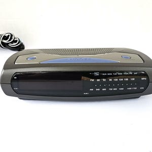 May include: A black and silver clock radio with a digital display and a blue accent. The clock radio has a built-in alarm and a radio tuner. The clock radio has a battery low indicator and a day of the week display. The clock radio has an AM/FM radio tuner with a frequency range of 88-108 MHz and 540-1700 kHz.