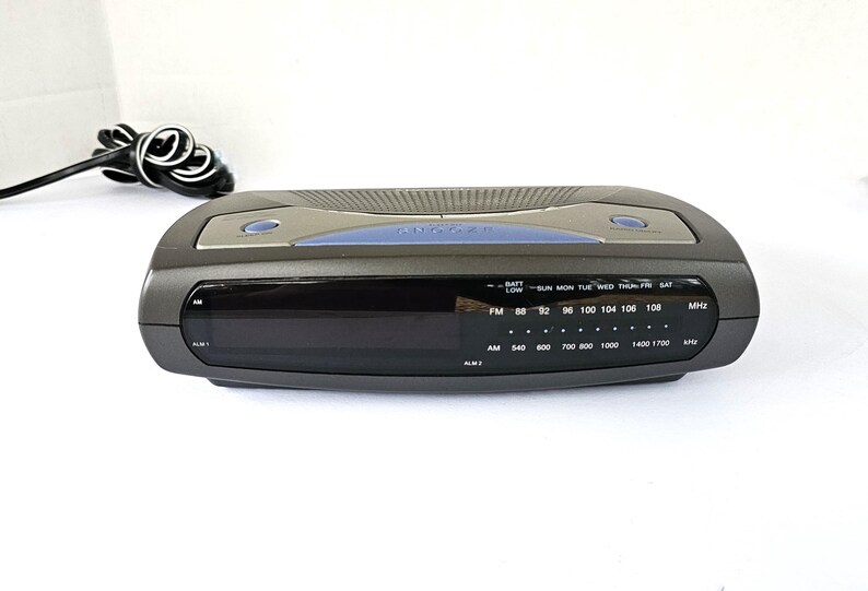 May include: A black and silver clock radio with a digital display and a blue accent. The clock radio has a built-in alarm and a radio tuner. The clock radio has a battery low indicator and a day of the week display. The clock radio has an AM/FM radio tuner with a frequency range of 88-108 MHz and 540-1700 kHz.