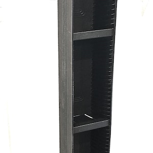 May include: A tall, black, vertical media storage tower featuring a rectangular base and multiple internal slots designed for organizing discs. The unit has a textured exterior finish and two fixed horizontal shelves dividing the interior space.