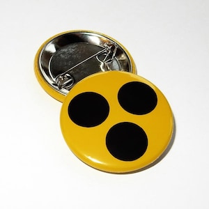 Może przedstawiać: Dwa żółte, okrągłe guziki z czarnym wzorem z trzema kropkami. Jeden guzik jest skierowany w górę, pokazując wzór, a drugi jest skierowany w dół, odsłaniając agrafkę. Guziki są na białym tle.