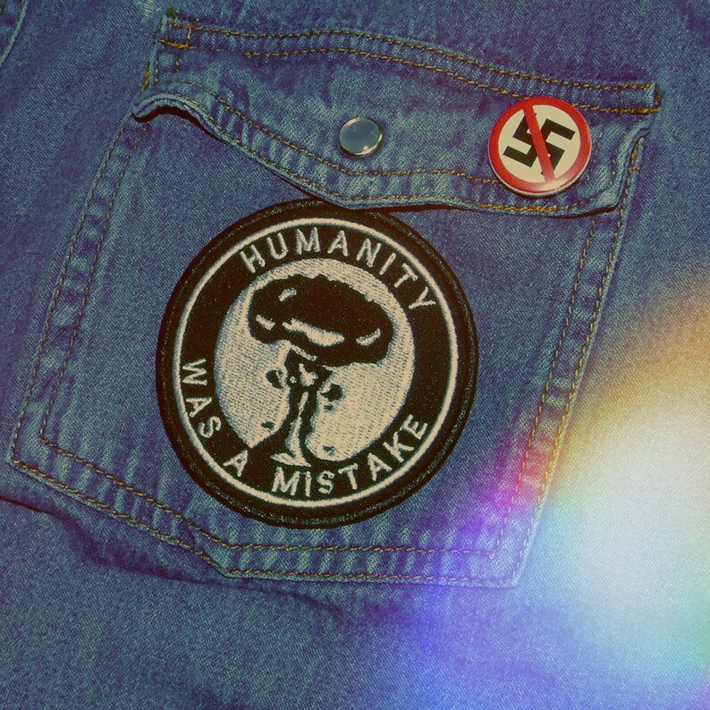 Może przedstawiać: Niebieska dżinsowa kurtka z czarną naszywką z napisem "HUMANITY WAS A MISTAKE" i czerwonym k&oacute;łkiem z czarnym symbolem w środku. Symbol to koło z przekątną linią i mniejszym k&oacute;łkiem wewnątrz większego koła.