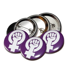 May include: A collection of purple and white feminist buttons. The buttons feature a white fist inside a white Venus symbol on a purple background. The backs of the buttons are silver with a pin closure.