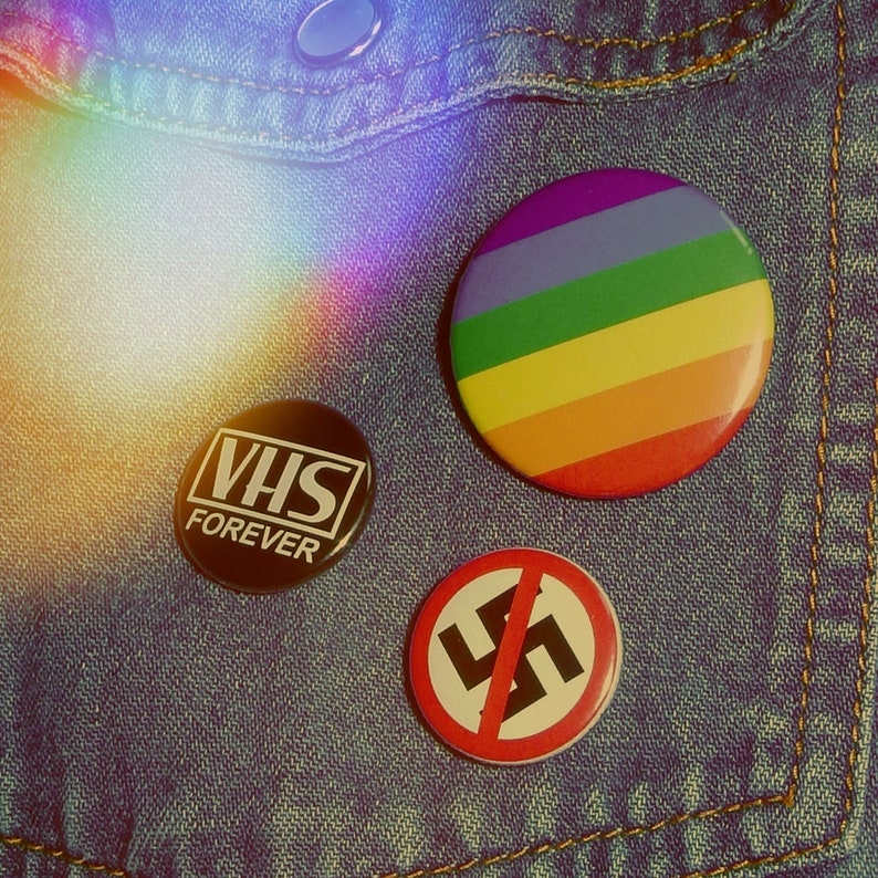 Może przedstawiać: Trzy odznaki na denimowym tle. Odznaki są okrągłe i mają r&oacute;żne wzory. Jedna odznaka jest czarna z białym tekstem, kt&oacute;ry brzmi "VHS FOREVER". Inna odznaka jest tęczowa z wzorem tęczowych pask&oacute;w. Trzecia odznaka jest czerwona z czarnym symbolem swastyki i tekstem "Nie" w kolorze czarnym.