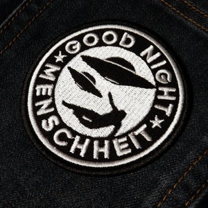 Może przedstawiać: Naszywka haftowana w kolorze czarnym i białym z tekstem "Good Night Menschheit" w k&oacute;łku.