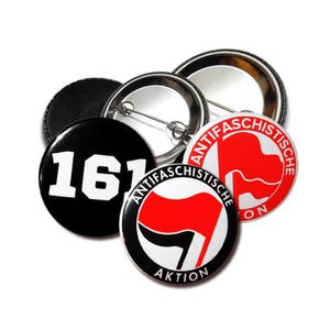 Puede incluir: Un conjunto de botones redondos con varios diseños. Un botón es negro con el número "161" en blanco. Otros botones presentan las palabras "ANTIFASCHISTISCHE AKTION" y un diseño de bandera roja sobre fondo blanco.
