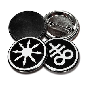 Op de afbeelding: Drie zwart-witte knoopsluitingen met verschillende ontwerpen. De eerste knoopsluiting heeft een witte zon met acht witte pijlen die naar buiten wijzen. De tweede knoopsluiting heeft een wit symbool met een kruis en een oneindigheidssymbool. De derde knoopsluiting is leeg.