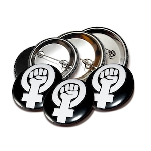 Könnte beinhalten: Drei schwarze und weiße feministische Faust-Knöpfe mit silberner Rückseite. Die Knöpfe zeigen eine weiße Faust in einem Kreis mit einem weißen weiblichen Symbol.