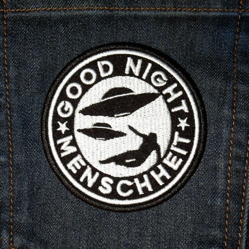 Może przedstawiać: Naszywka haftowana w kolorze czarnym i białym z tekstem "Good Night Menschheit" w k&oacute;łku. Naszywka przedstawia trzy latające spodki i sylwetkę osoby.