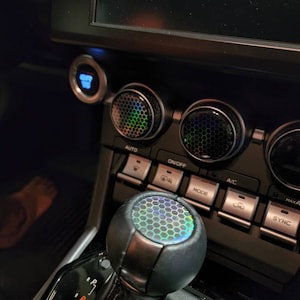 Puede incluir: Una palanca de cambios de coche de cuero negro con un patrón hexagonal iridiscente en la parte superior. La palanca está rodeada de un borde plateado y está unida a una funda de palanca de cambios de cuero negro.