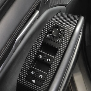 May include: A car door panel with carbon fibre trim around the window controls. The panel has a black control panel with window and lock buttons, and a circular control dial. The door panel is black with stitched leather detailing.