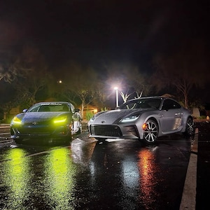 Puede incluir: Dos coches deportivos plateados aparcados en un aparcamiento mojado por la noche. El coche de la izquierda tiene una pegatina amarilla en el parabrisas que dice "FIRE RACK".