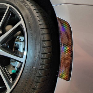 May include: A close-up of a silver car with a black tire and a black and iridescent honeycomb-shaped sticker on the fender.