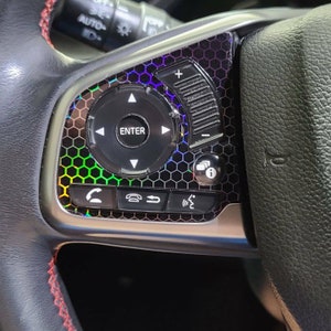 Può includere: Primo piano di un volante di un'auto con un motivo a nido d'ape nero e iridescente. Il volante ha controlli per il volume, le chiamate telefoniche e la navigazione. I controlli sono etichettati con icone e la parola "ENTER".