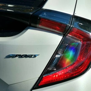 May include: A white car with a red rear light and the word "SPORT" in silver lettering on the back. The rear light has a rainbow iridescent film over the lens.