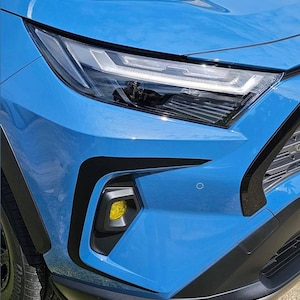 K&ouml;nnte beinhalten: Ein blaues Auto mit schwarzen Zierleisten und einem gelben Blinklicht. Das Auto ist auf einer asphaltierten Fl&auml;che geparkt.