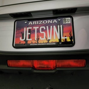 Puede incluir: Un coche plateado con una matrícula negra que dice "ARIZONA" y "JETSVNL". La matrícula tiene un diseño de puesta de sol y cactus. El coche tiene una luz trasera roja.