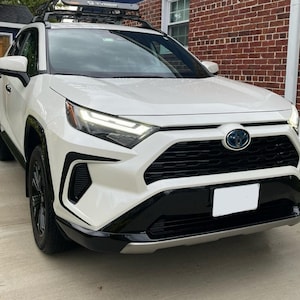 K&ouml;nnte beinhalten: Ein wei&szlig;er Toyota RAV4 mit schwarzen Akzenten, darunter ein schwarzer K&uuml;hlergrill, schwarze Scheinwerfer und schwarze Felgen. Das Auto hat ein Dachgep&auml;cktr&auml;ger und ein Nummernschild.