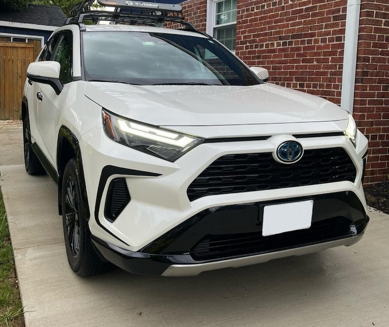 K&ouml;nnte beinhalten: Ein wei&szlig;er Toyota RAV4 mit schwarzen Akzenten, darunter ein schwarzer K&uuml;hlergrill, schwarze Scheinwerfer und schwarze Felgen. Das Auto hat ein Dachgep&auml;cktr&auml;ger und ein Nummernschild.