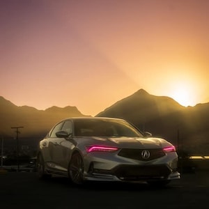 Puede incluir: Un sedán plateado con faros rosados estacionado frente a una cordillera al atardecer.