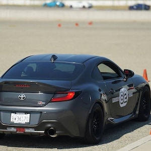 Puede incluir: Un coche deportivo Toyota GR86 gris oscuro con un alerón trasero negro y un difusor trasero negro. El coche tiene una franja de carreras negra en el lateral y un número blanco "82" en el lateral. El coche está en una superficie de asfalto gris con conos naranjas.