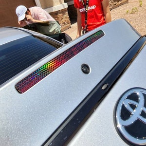 Puede incluir: Un coche plateado con un parachoques trasero negro y un emblema de Toyota negro y plateado. El coche tiene una pegatina iridiscente arcoíris en la luneta trasera. La pegatina tiene un patrón de panal.
