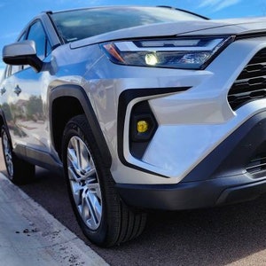 K&ouml;nnte beinhalten: Ein silberner Toyota RAV4 mit einem schwarzen K&uuml;hlergrill und gelben Nebelscheinwerfern. Das Auto ist auf einer asphaltierten Stra&szlig;e mit einem B&uuml;rgersteig und gr&uuml;nen B&uuml;schen im Hintergrund geparkt.