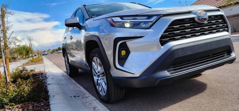 K&ouml;nnte beinhalten: Ein silberner Toyota RAV4 mit einem schwarzen K&uuml;hlergrill und gelben Nebelscheinwerfern. Das Auto ist auf einer asphaltierten Stra&szlig;e mit einem B&uuml;rgersteig und gr&uuml;nen B&uuml;schen im Hintergrund geparkt.
