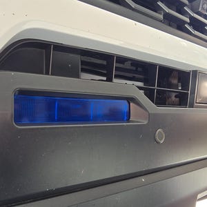 May include: A white car with a black front bumper and a blue light bar. The car has a black grille with a silver trim. The car is parked on a driveway.