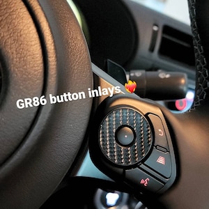 May include: Close-up of a black steering wheel with a carbon fiber button inlay. The button has a black center button with a red ring around it. The text "GR86 button inlays" is written in white on the steering wheel.