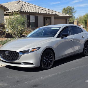 Puede incluir: Un sedán Mazda 3 gris plateado estacionado frente a una casa. El coche tiene llantas negras y ventanas tintadas. El coche está estacionado en una entrada pavimentada.