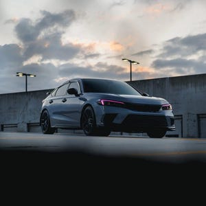 May include: A gray sedan with black wheels and accents is parked on a concrete surface. The car has pink lights and is set against a cloudy sky. The image is taken at a low angle.