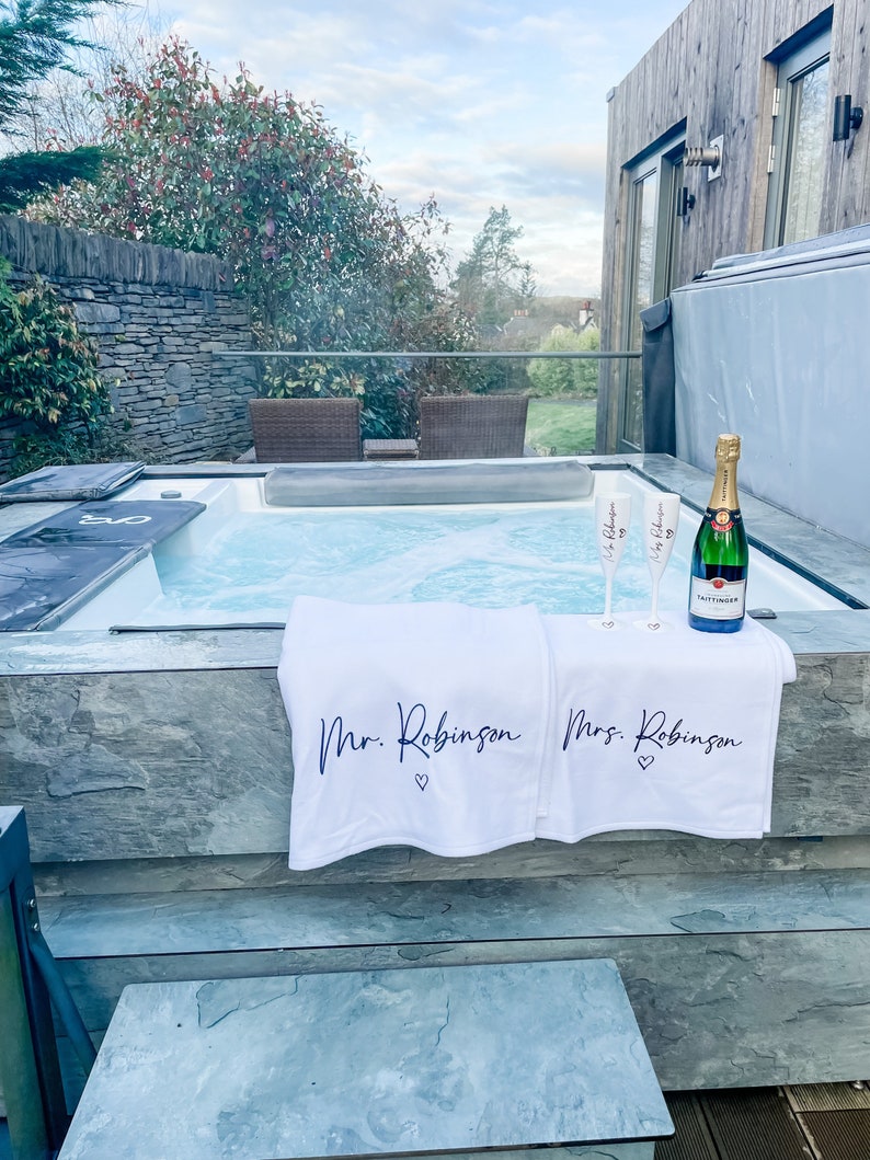 May include: Two white towels with the text "Mr. Robinson" and "Mrs. Robinson" on them, placed on the edge of a hot tub. Two champagne flutes and a bottle of champagne are in the hot tub.