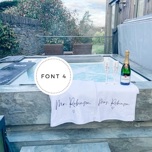 May include: Two white towels with the text "Mr. Robinson" and "Mrs. Robinson" with a heart symbol on each towel. The towels are hanging on the edge of a hot tub with two champagne flutes and a bottle of champagne.