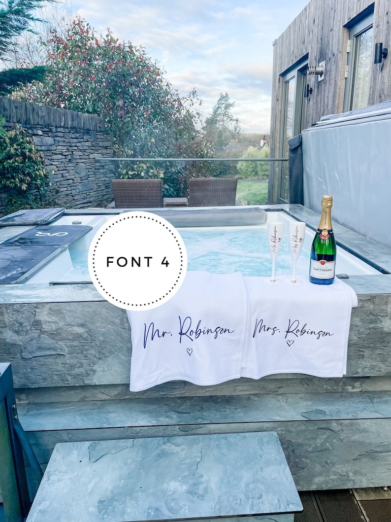 May include: Two white towels with the text "Mr. Robinson" and "Mrs. Robinson" with a heart symbol on each towel. The towels are hanging on the edge of a hot tub with two champagne flutes and a bottle of champagne.