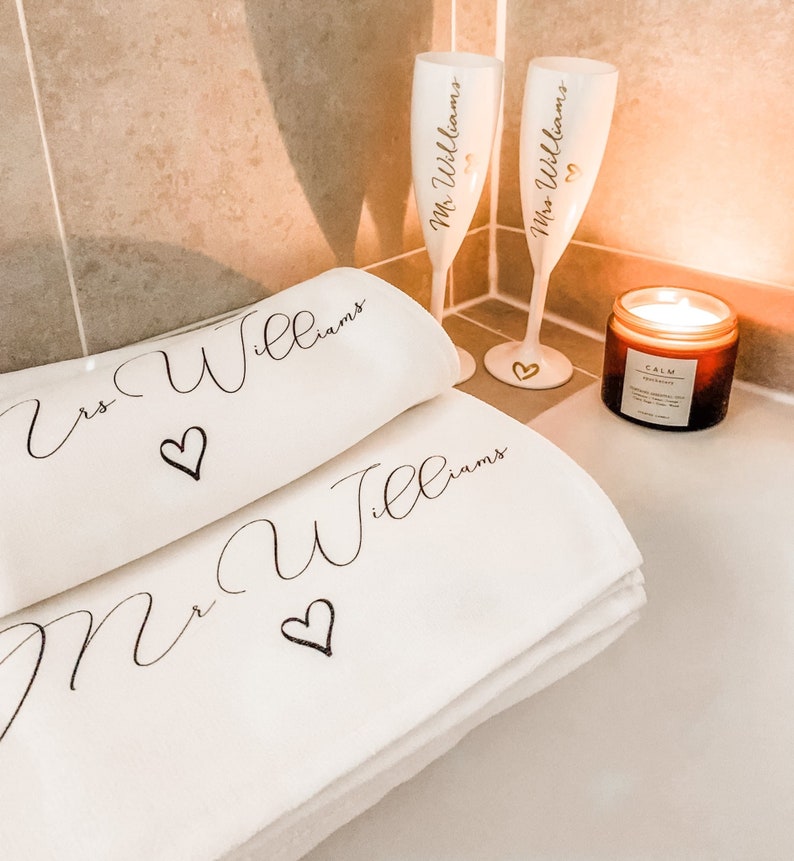 May include: Two white champagne flutes with gold lettering that says "Mrs. Williams" and "Mr. Williams" with a heart symbol. The flutes are on a white surface with two white towels that say "Mrs. Williams" and "Mr. Williams" with a heart symbol. There is a brown candle with a white label that says "Calm" on the surface.