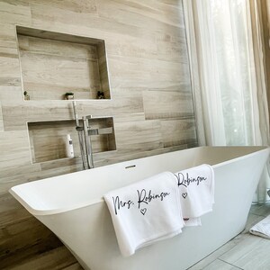 May include: A white bathtub with two white towels draped over the edge. The towels have black lettering that says "Mrs. Robinson" and "Robinson" with a small heart symbol next to each name.