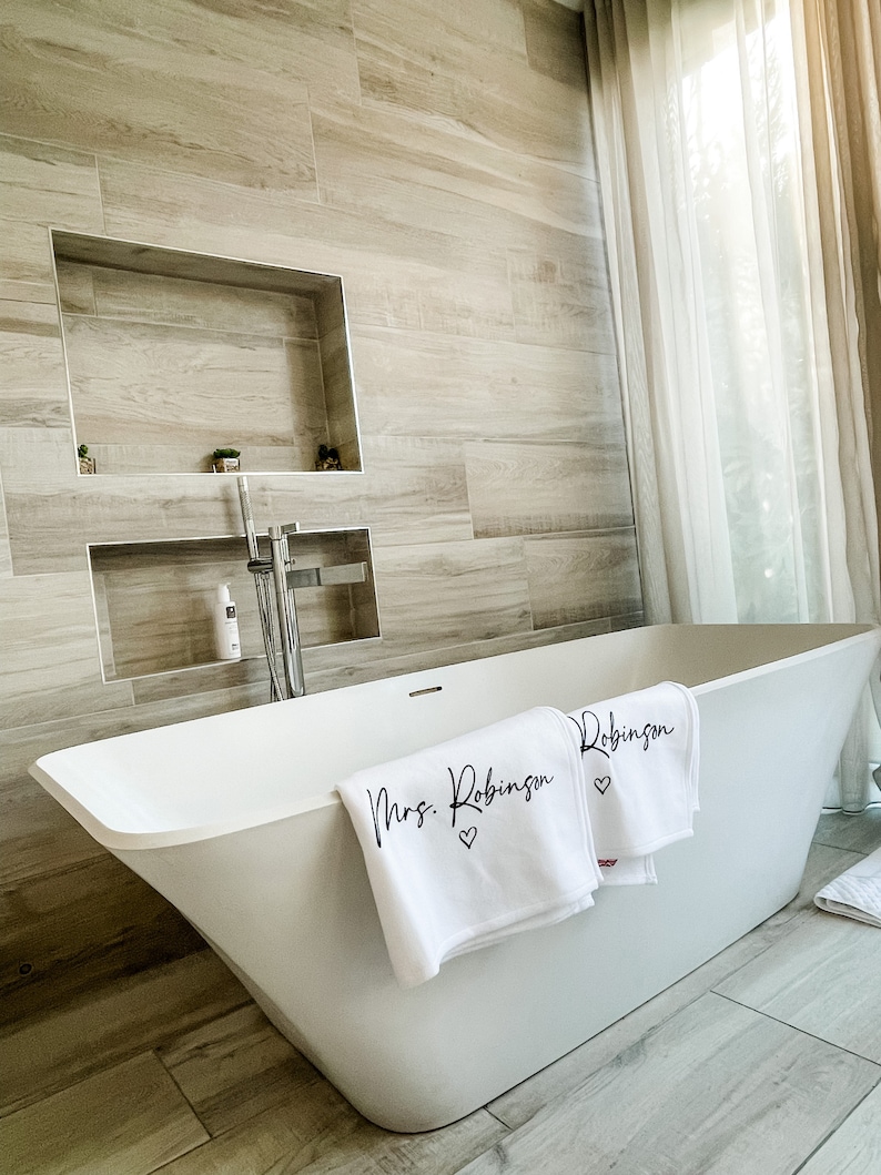 May include: A white bathtub with two white towels draped over the edge. The towels have black lettering that says "Mrs. Robinson" and "Robinson" with a small heart symbol next to each name.