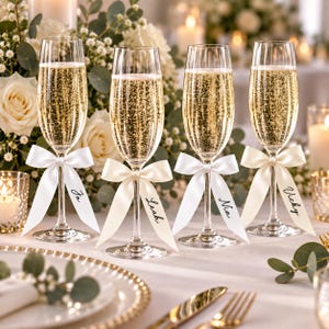 May include: Four clear glass champagne flutes filled with sparkling liquid, each featuring a white satin ribbon tied around the stem. The ribbons are printed with the names Jo, Leah, Nia, and Vicky in a black cursive script font.