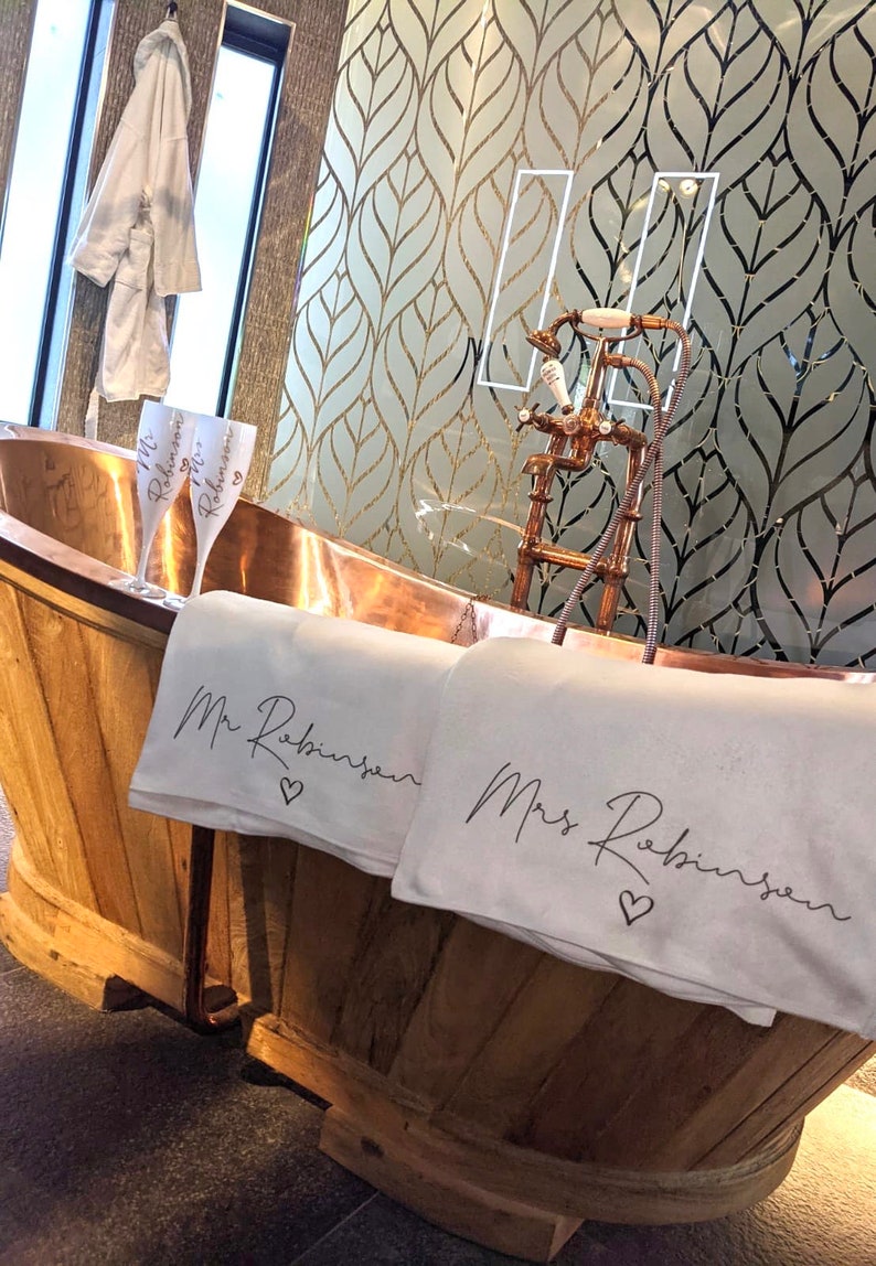 May include: A copper bathtub with two white towels, one with "Mr. Robinson" and the other with "Mrs. Robinson" embroidered on them. Two champagne flutes with "Mr. Robinson" and "Mrs. Robinson" on them are in the tub.