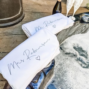 May include: Two white towels with the text "Mr. Robinson" and "Mrs. Robinson" in black script with a heart symbol. The towels are folded and placed on a wooden surface next to a hot tub. Two champagne flutes with the text "Mr. Robinson" and "Mrs. Robinson" are on the edge of the hot tub.