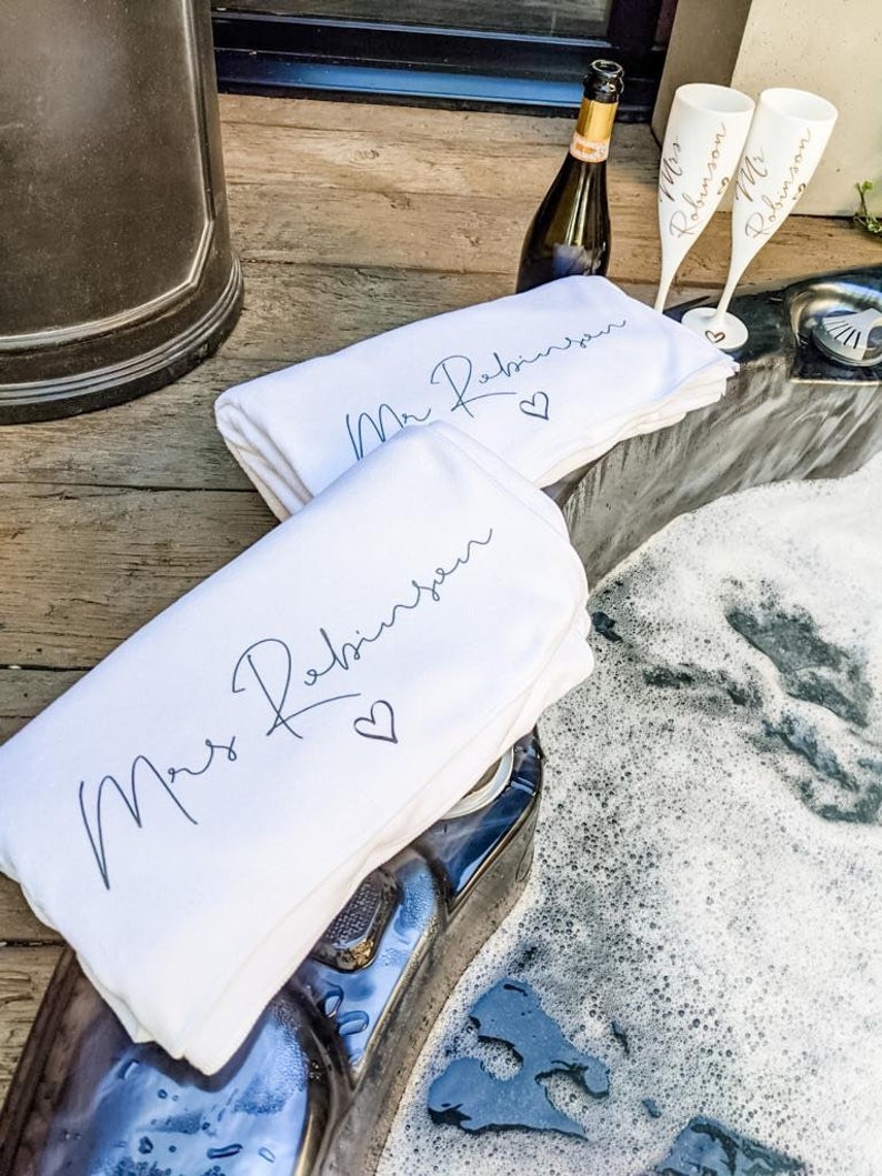May include: Two white towels with the text "Mr. Robinson" and "Mrs. Robinson" in black script with a heart symbol. The towels are folded and placed on a wooden surface next to a hot tub. Two champagne flutes with the text "Mr. Robinson" and "Mrs. Robinson" are on the edge of the hot tub.